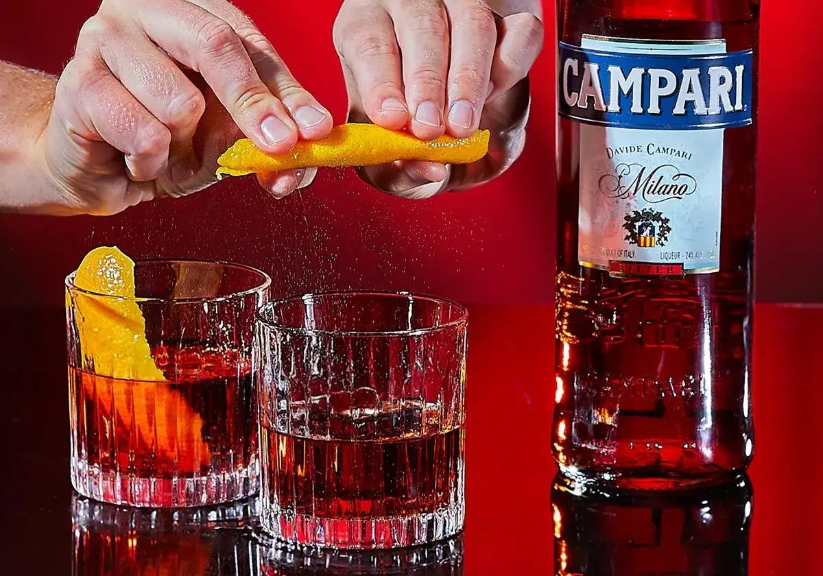 Przygotowanie drinka Negroni: ręka wyciska skórkę pomarańczy nad szklanką z czerwonym alkoholem i lodem. Obok butelka Campari.