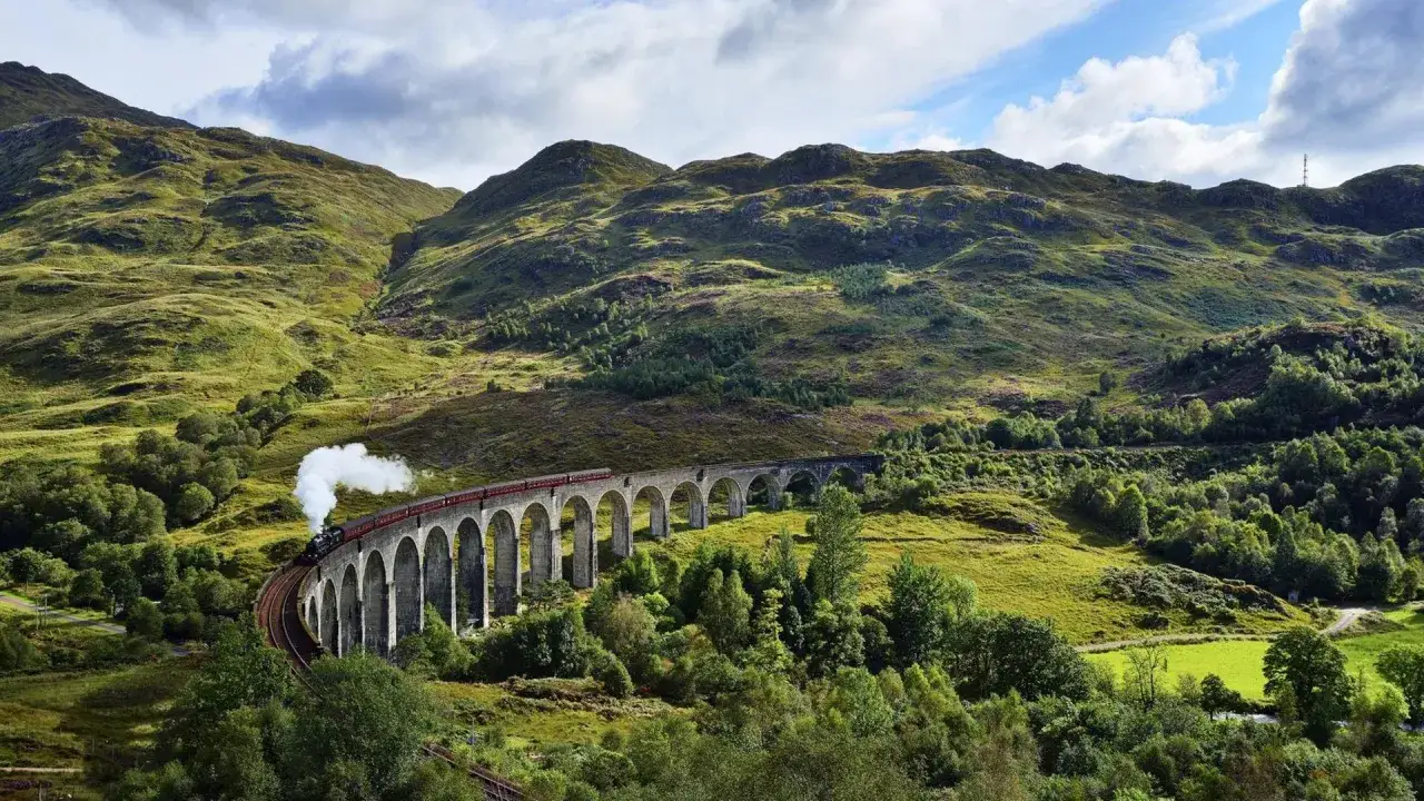 Dampfender Zug fährt über eine Bogenbrücke in den schottischen Highlands, umgeben von grünen Hügeln und Bäumen.