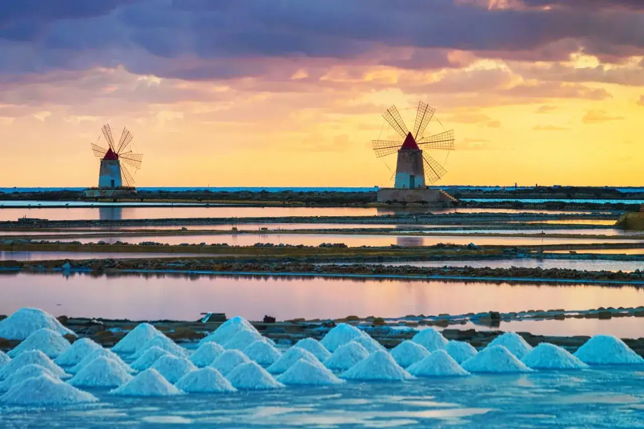 Saline di Trapani al tramonto, con mulini a vento e cumuli di sale. Un'idea per cosa vedere a Trapani in un giorno.