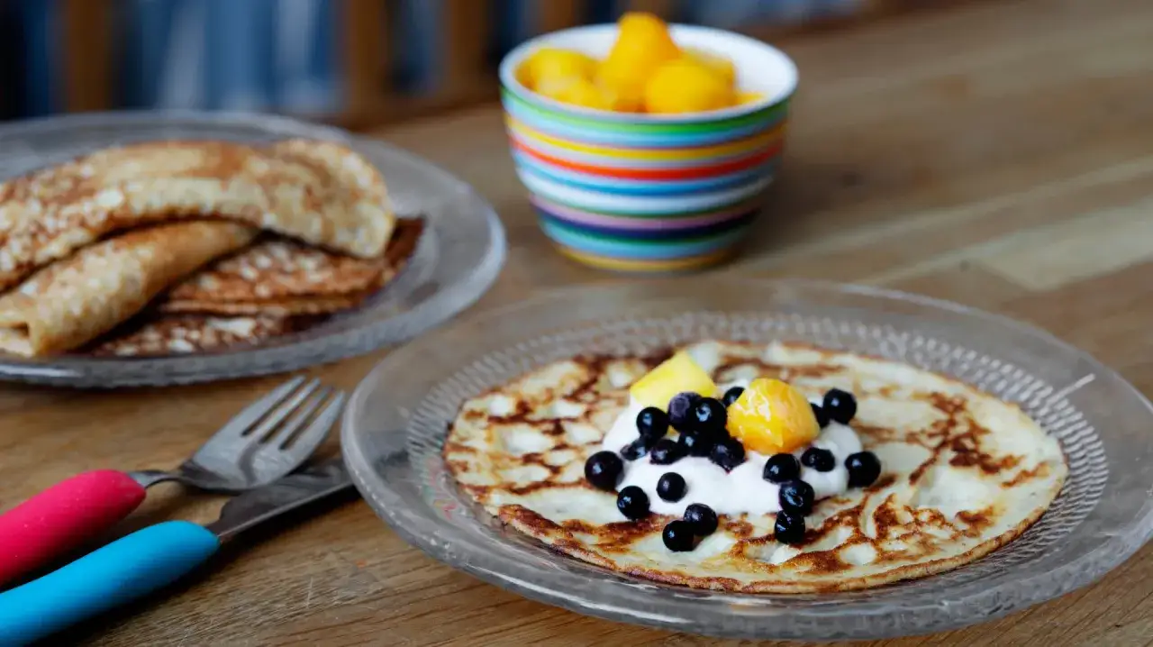 En tallrik med pannkakor med havregryn, toppade med grädde, blåbär och frukt. En annan tallrik med pannkakor syns i bakgrunden.