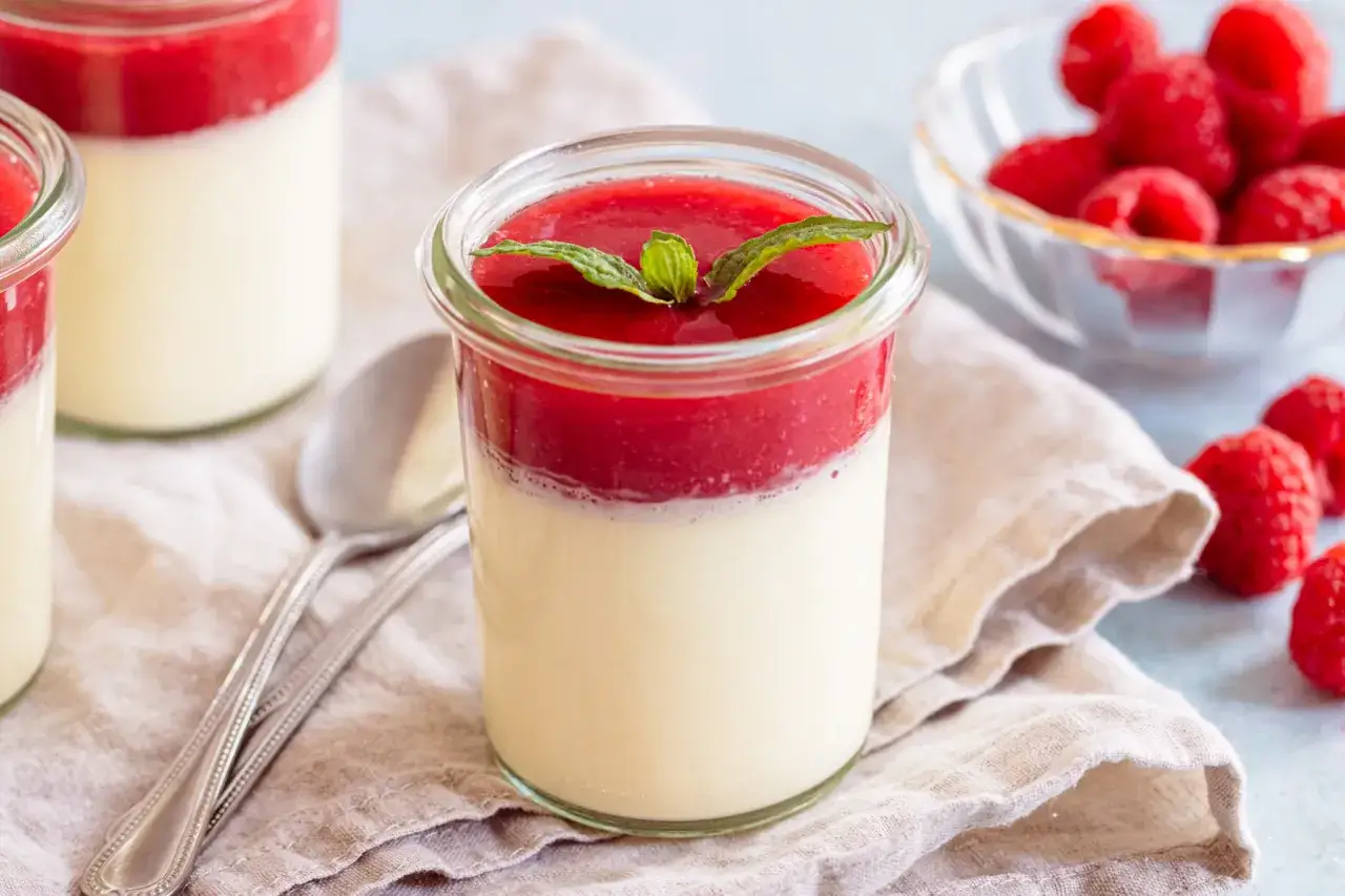 Vegane Panna Cotta mit Himbeersoße und Minze, serviert in einem Glas. Daneben frische Himbeeren und ein Löffel.
