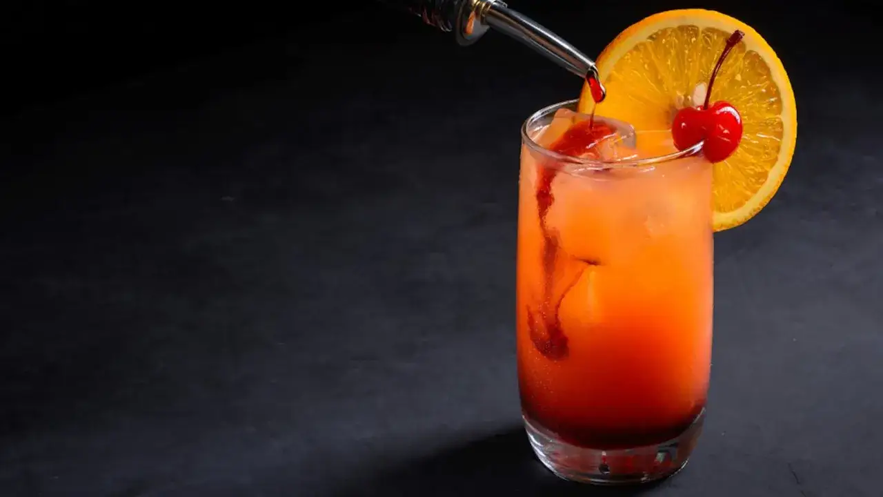 Kolorowy drink z grenadyną, ozdobiony plasterkiem pomarańczy i wisienką, z ciemnym tłem.