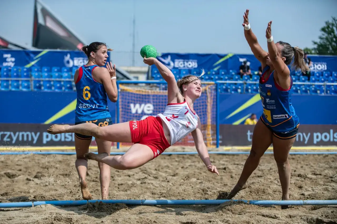 Piłkarka w skoku po zieloną piłkę podczas meczu beach handball. Rywalka w niebieskiej koszulce z numerem 6 obserwuje akcję.