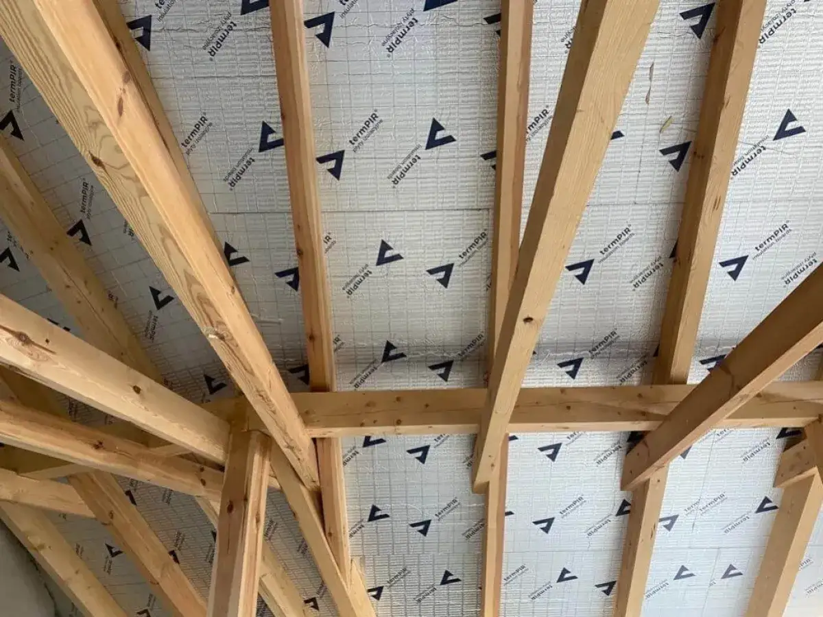 Drewniana konstrukcja dachu z widocznym ociepleniem ściany szczytowej od góry. Materiał izolacyjny z logo