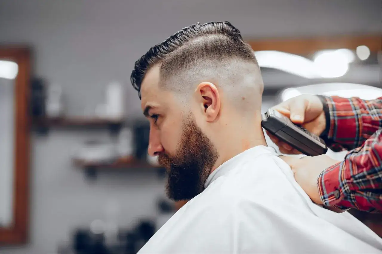 Un homme avec un **modèle barbe dégradé** se fait raser la nuque par un barbier en chemise à carreaux.