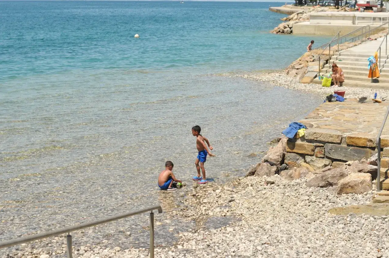 Zwei Jungen spielen am flachen, steinigen slowenien strand. Das klare Wasser l&auml;dt zum Planschen ein, w&auml;hrend im Hintergrund eine Frau auf den Stufen sitzt.
