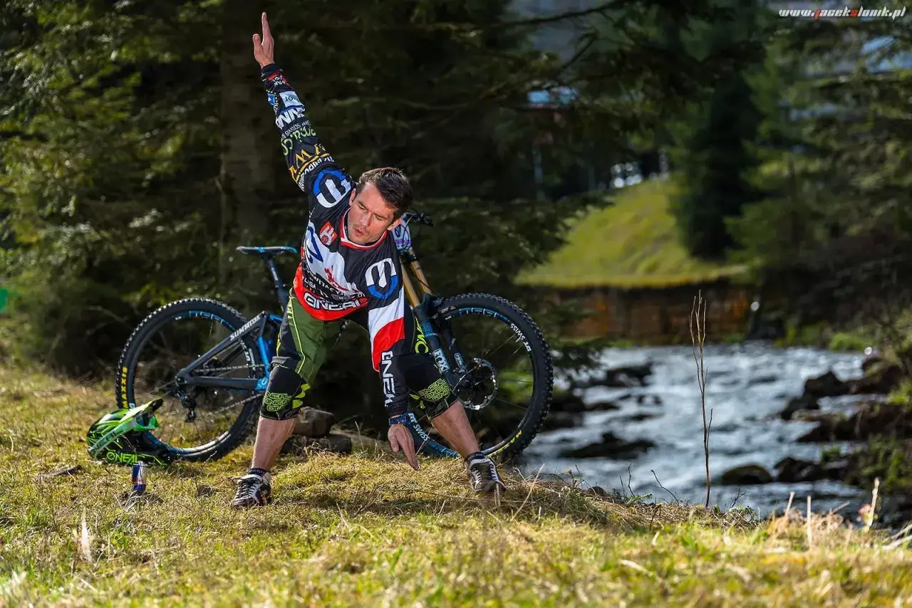 Kolarz w stroju z logo O'Neal wykonuje ćwiczenia na rozgrzewkę przed zjazdem. Obok rower i kask.