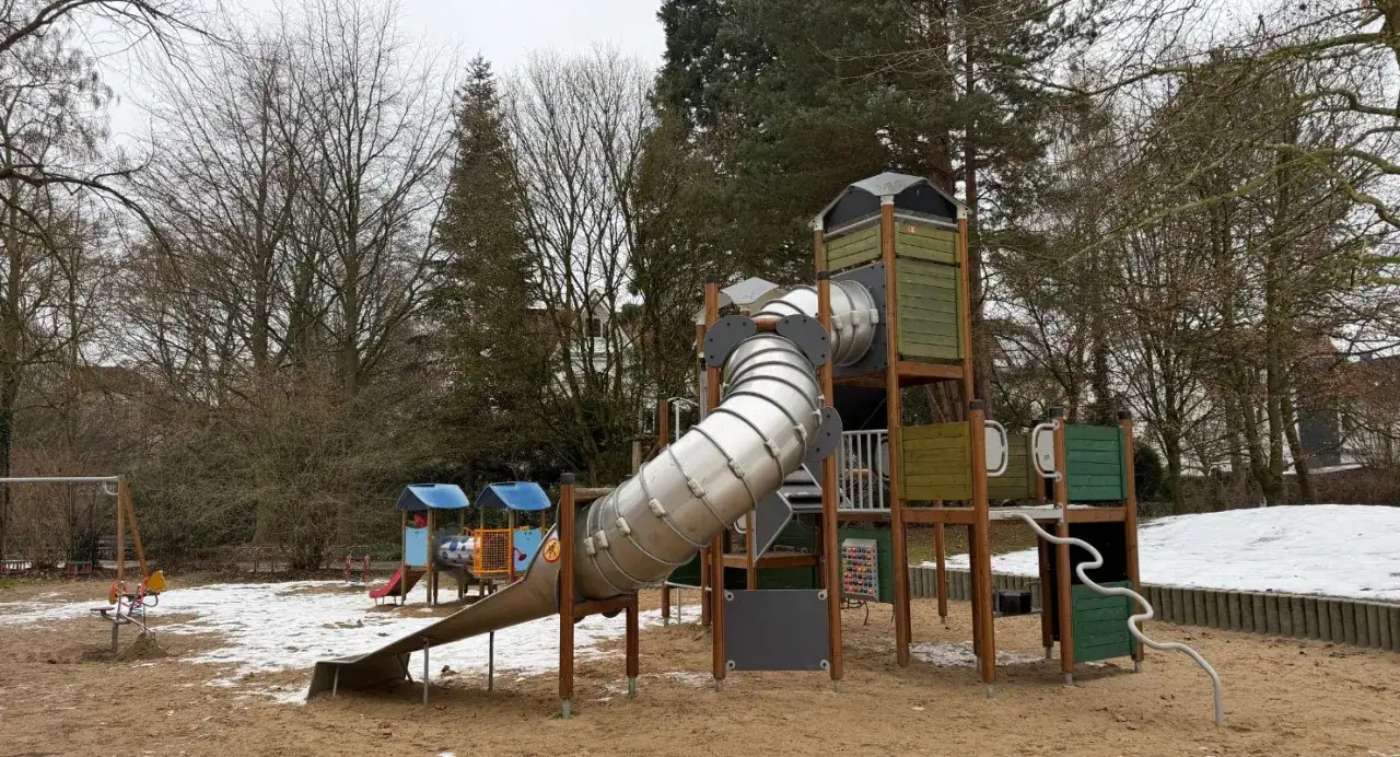 Ein Spielplatz im Herford Park mit einer großen Röhrenrutsche und Schaukeln, umgeben von kahlen Bäumen und etwas Schnee.