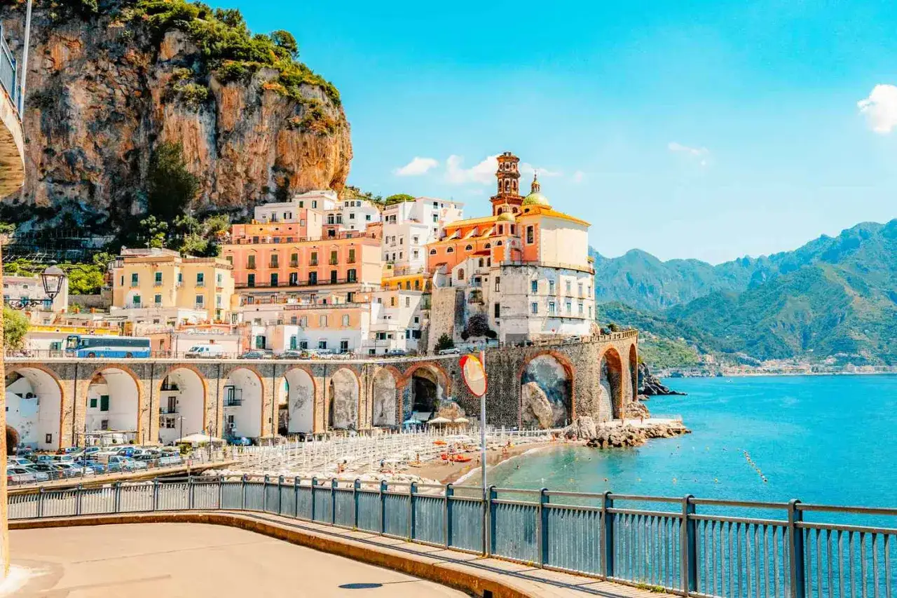 F&auml;rgstarka hus kl&auml;ttrar uppf&ouml;r klippan vid havet, ett dr&ouml;m **italien resm&aring;l**. En strand med parasoller och en kyrka med kupol.