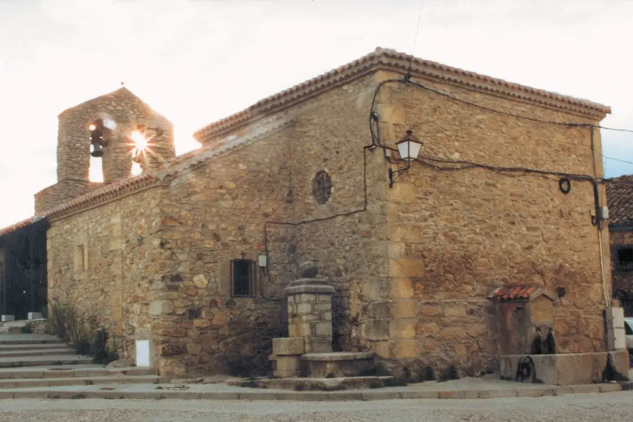 Iglesia de piedra con campanario y sol brillando. Una fuente antigua adorna la plaza de la acebeda.