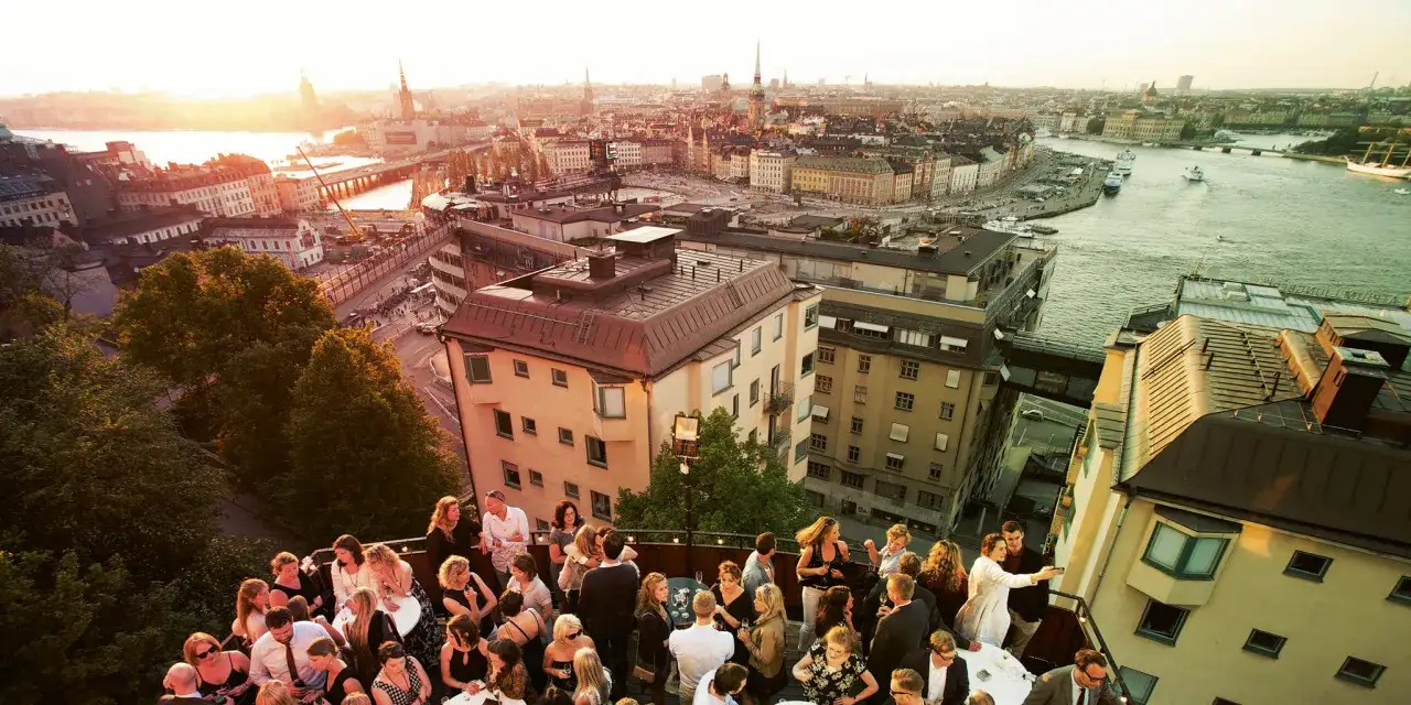 Festlig sammankomst på en takterrass med utsikt över Stockholm. Solnedgången lyser upp staden och vattnet. Södra teatern skymtar i bakgrunden.