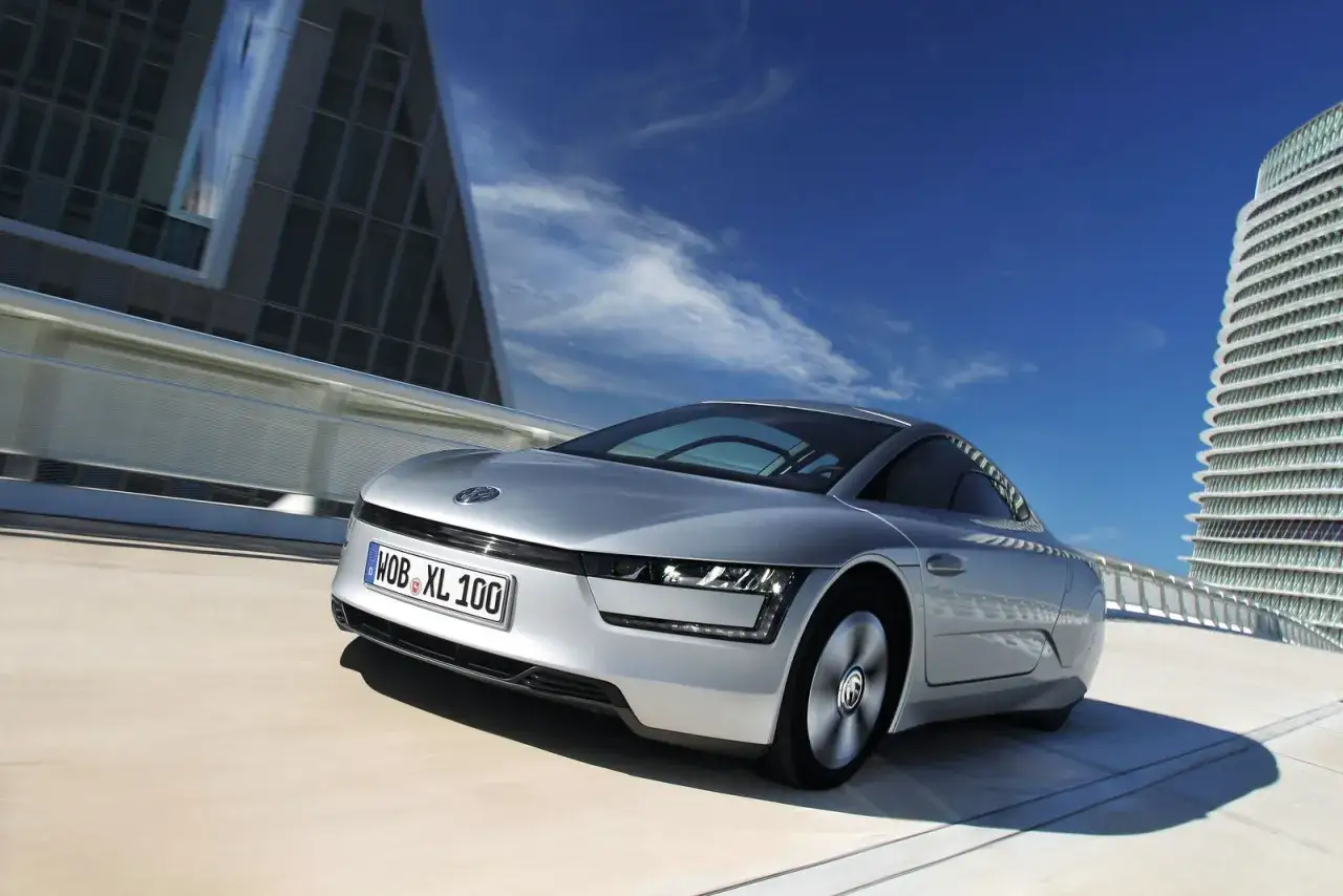 Srebrny VW XL1 pędzi po nowoczesnej drodze, otoczony futurystyczną architekturą i błękitnym niebem.