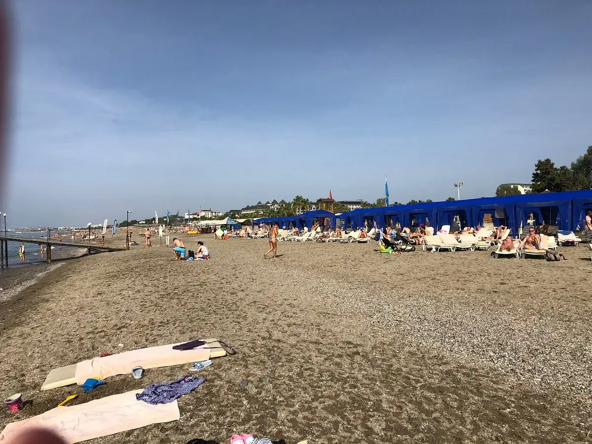 Plaża z leżakami pod niebieskimi namiotami. Ludzie odpoczywają, widać molo. Idealne miejsce na wakacje, warto sprawdzić club kastalia opinie.