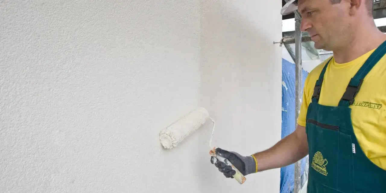 Pracownik w zielonym ogrodniczkach odnawia tynk strukturalny wałkiem.