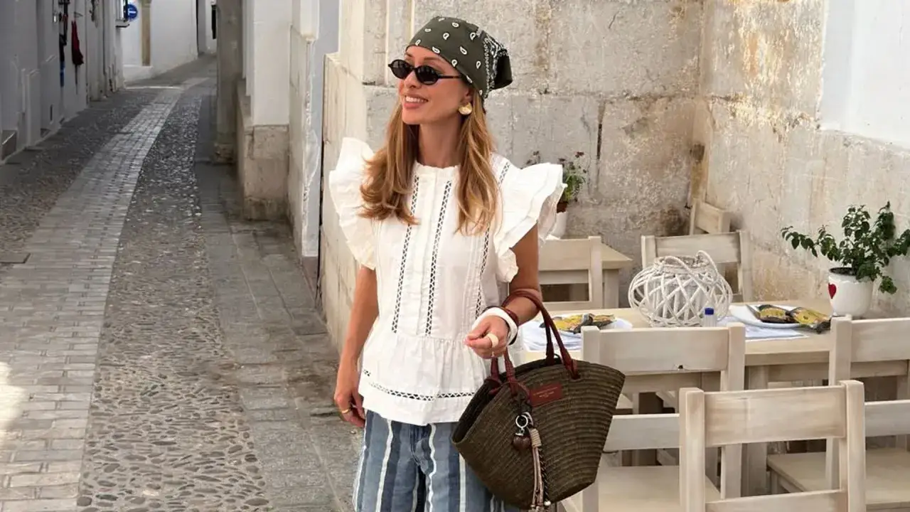 Mujer con pañuelo y gafas de sol, luciendo un top blanco y pantalones a rayas, en un callejón empedrado. Su bolso de mimbre complementa su look de street style.
