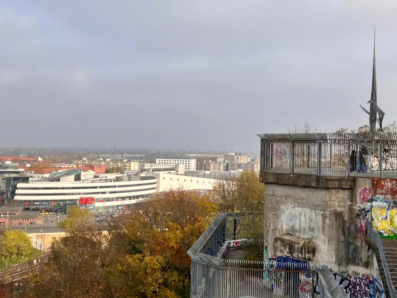 Blick vom Humboldthain Park &uuml;ber Berlin. Ein Denkmal ragt in den grauen Himmel, w&auml;hrend unten die Stadt und bunte Herbstb&auml;ume zu sehen sind.