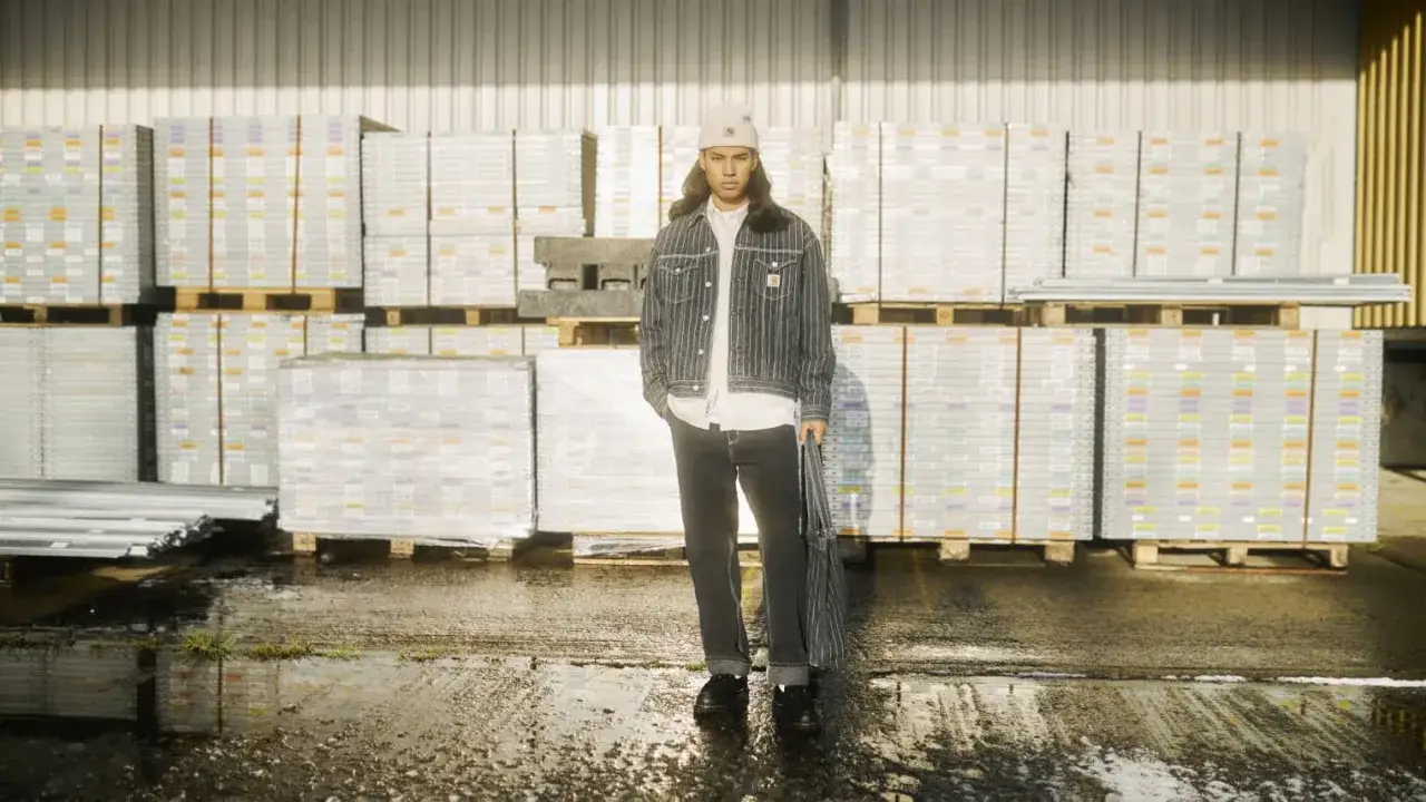 Młody mężczyzna w kurtce i spodniach Carhartt, stojący przed magazynem pełnym paczek. Carhartt to firma znana z odzieży roboczej.