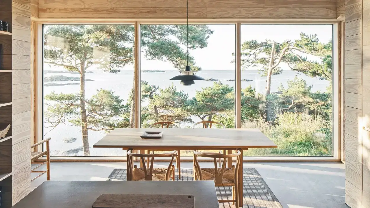 Interno di una casa stile norvegese con tavolo in legno e sedie, affacciata su un paesaggio marino con pini.