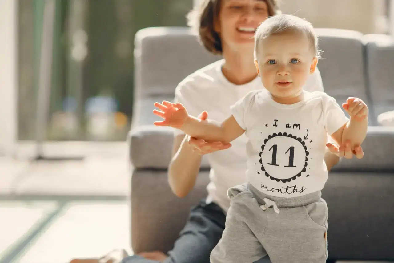Uśmiechnięta mama pomaga swojemu 11-miesięcznemu dziecku, kiedy dziecko siada, by zrobić pierwszy krok.