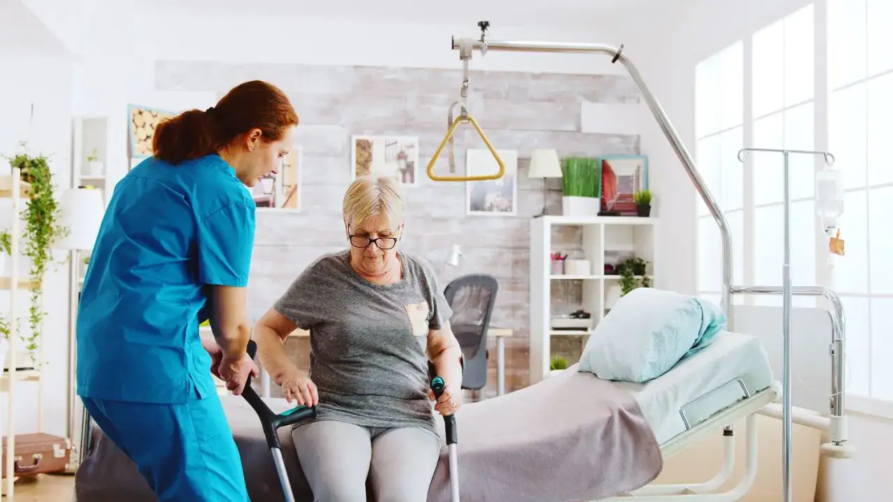 Une aide-soignante aide une femme âgée à se lever avec des béquilles, près d'un lit médicalisé gratuit.