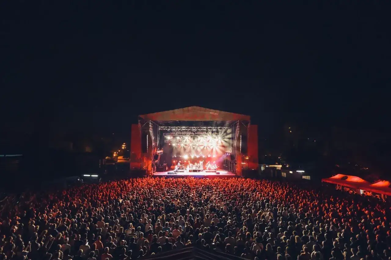 Multitud vibrante disfruta de los **concerts de Vivers** bajo un cielo nocturno, con una banda tocando en un escenario iluminado por luces rojas y blancas.