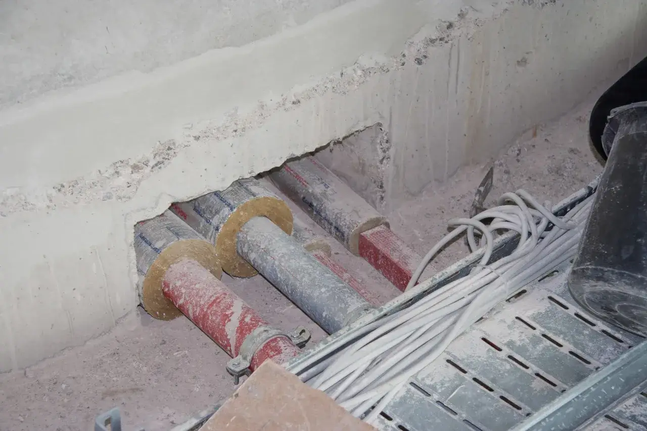 Rohre, isoliert nach DIN EN 13670, durchbrechen eine Betonwand. Kabel liegen auf einem Gittertr&auml;ger.