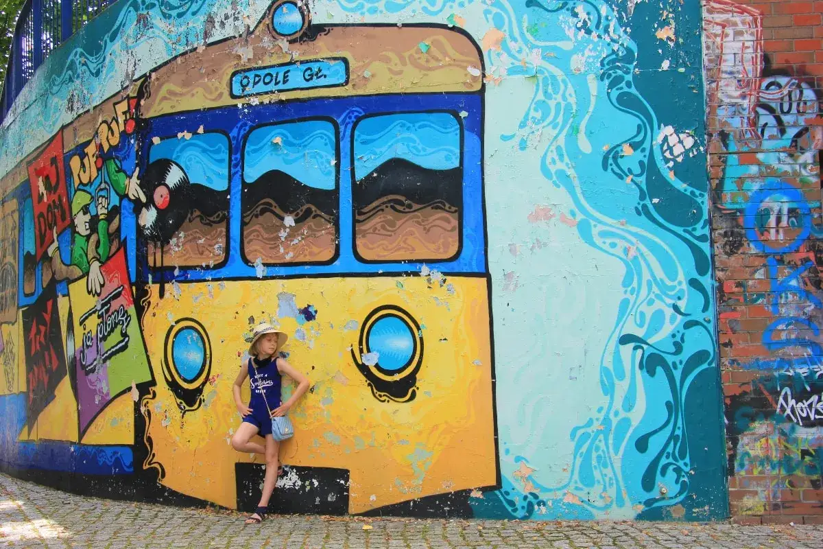 Dziewczynka w kapeluszu pozuje przy kolorowym muralu z tramwajem. To świetne opole atrakcje dla dzieci, pełne barw i zabawy.