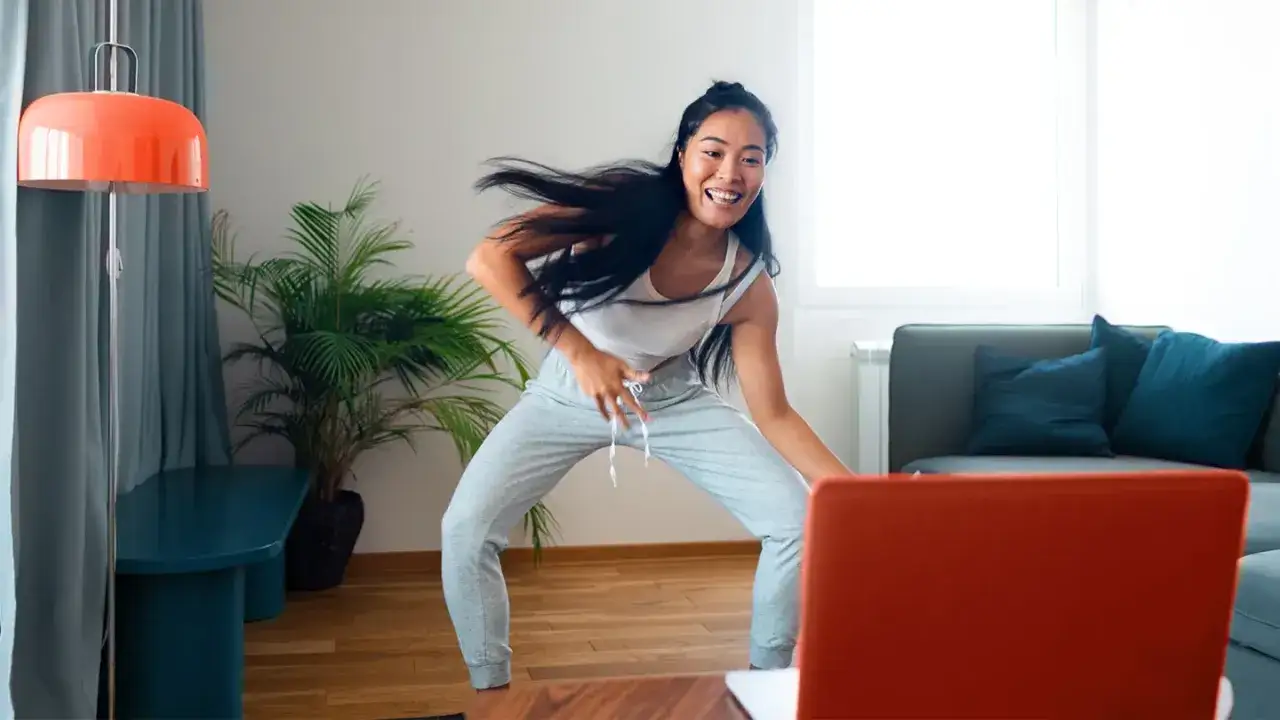 Uśmiechnięta kobieta ćwiczy zumbę w domu, tańcząc do muzyki z laptopa.