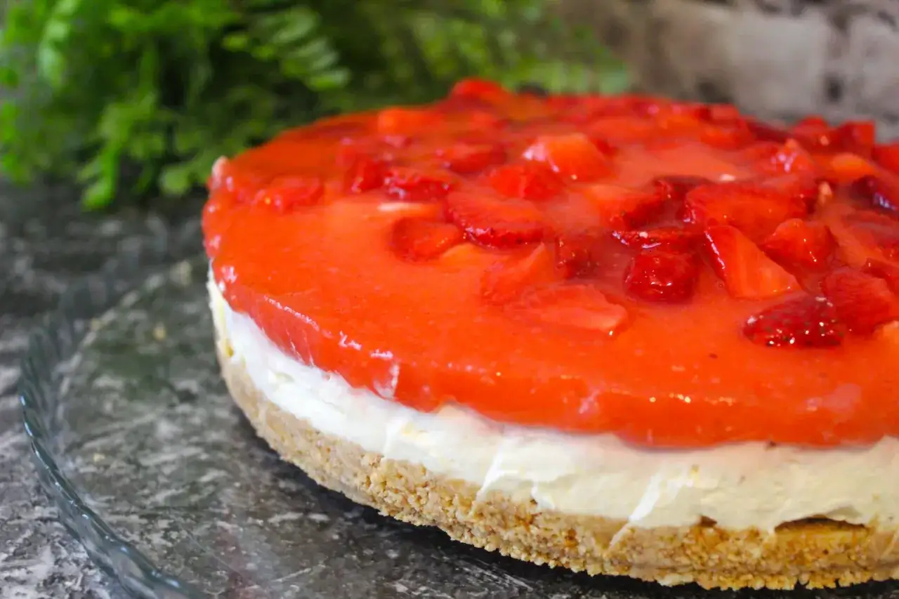 Torta fredda con base croccante, crema bianca e uno strato lucido di coulis di fragole e pezzi di frutta fresca.