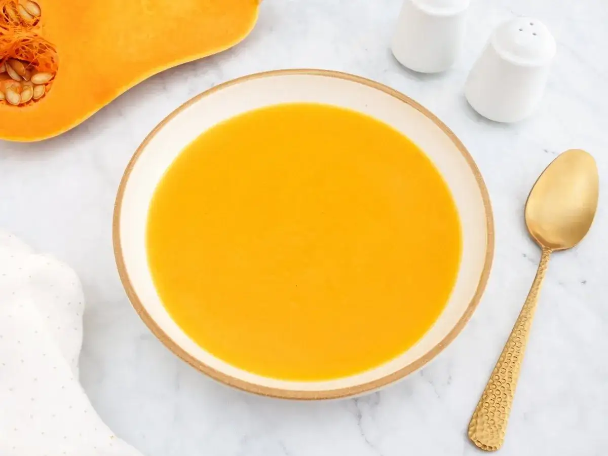 Un taz&oacute;n de cremoso **pure de calabaza** junto a una calabaza cortada, una cuchara dorada y saleros.