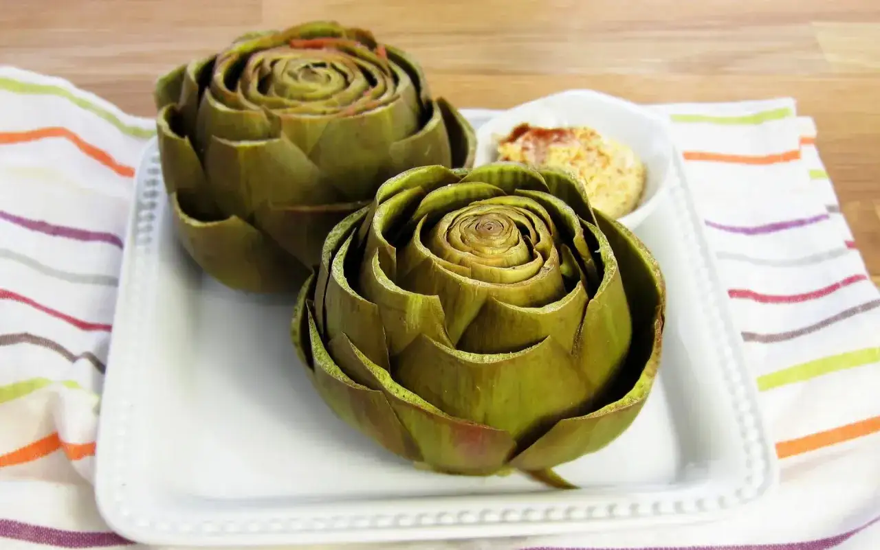Dos alcachofas listas para cocer, servidas en un plato blanco con un dip de hummus y especias.