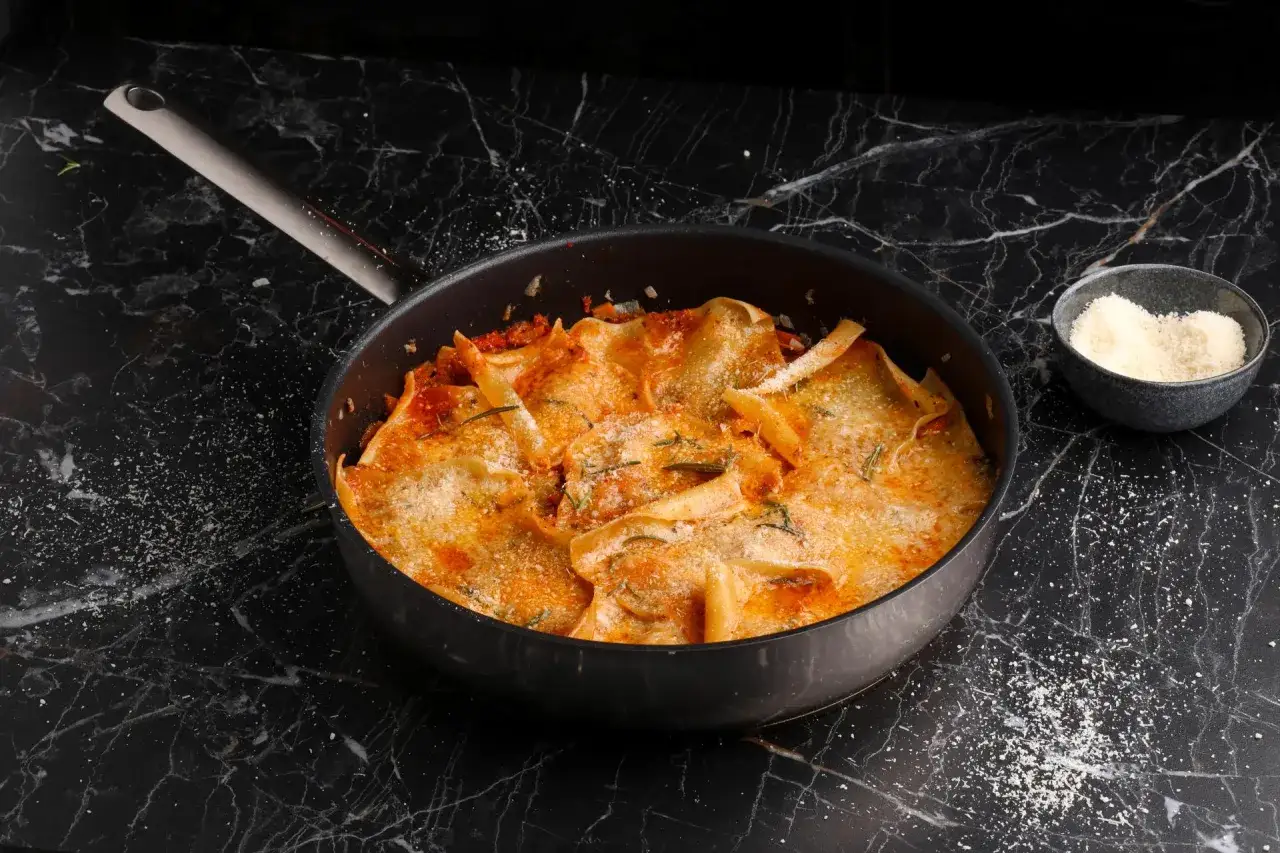 Herzhafte One Pot Lasagne in einer Pfanne, goldbraun &uuml;berbacken mit Parmesan und Rosmarin. Daneben eine Schale geriebenen K&auml;se.