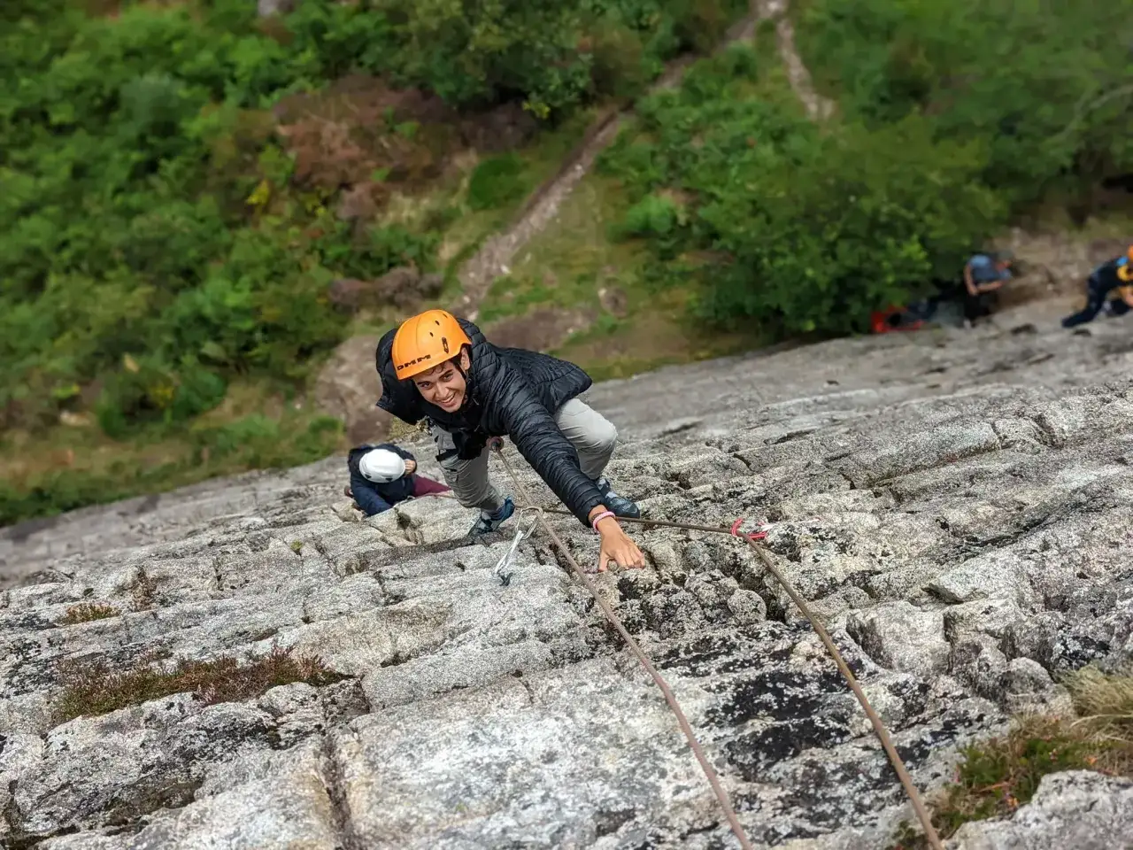 Un jeune grimpeur escalade une paroi rocheuse, souriant à l'objectif. D'autres personnes escaladent plus bas.