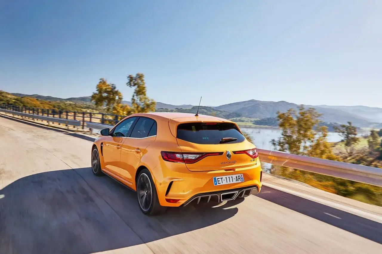 Pomarańczowy Renault Megane RS 4 pędzi po krętej drodze z widokiem na jezioro i wzg&oacute;rza.