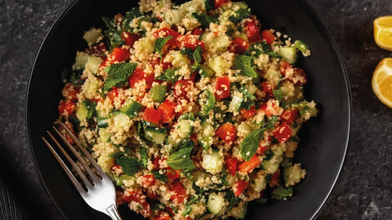 Frischer Couscous Salat mit Tomaten, Gurken und Minze in einer schwarzen Sch&uuml;ssel, daneben Zitronenspalten.
