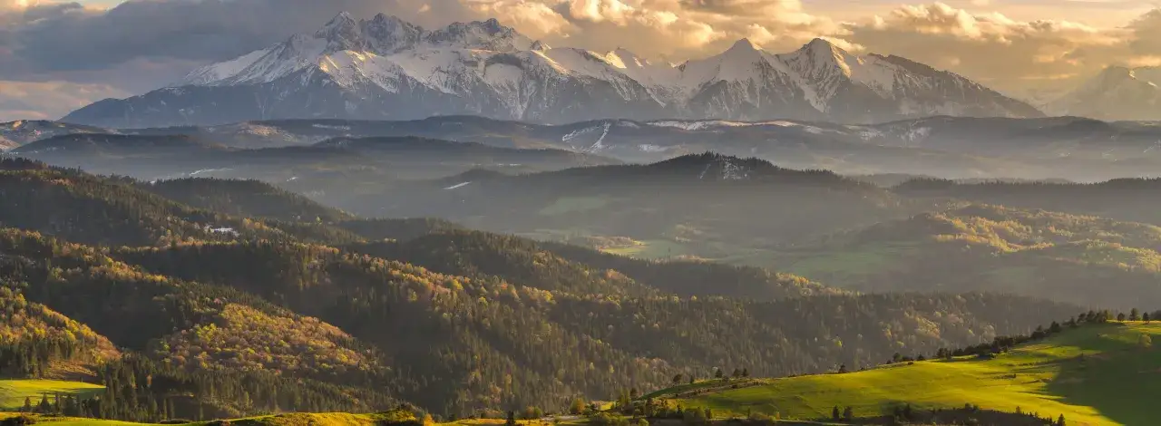 Widok na największe góry w Polsce, Tatry, ośnieżone szczyty w słońcu, na tle zielonych, zalesionych wzgórz.