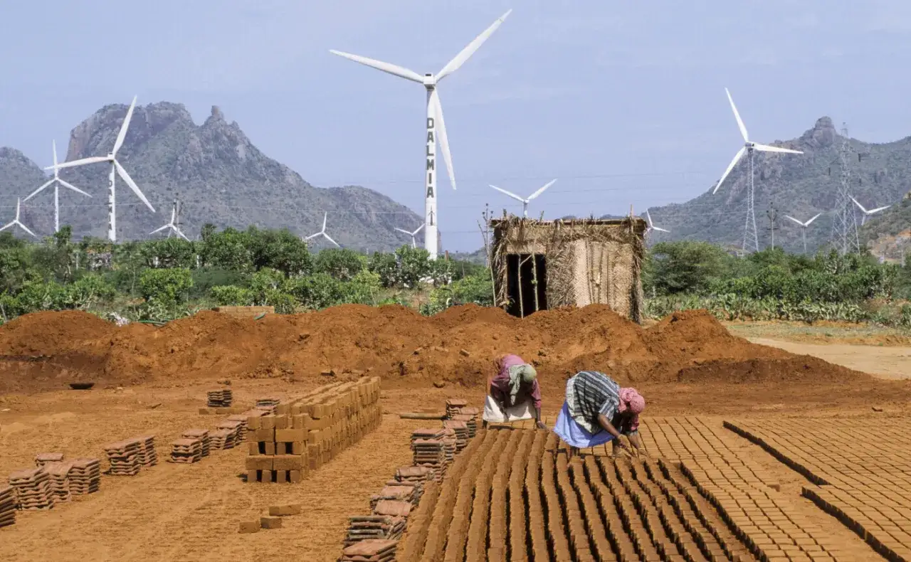 Zwei Frauen stellen Ziegel her, während im Hintergrund Windräder für den Klimaschutz in Indien stehen.