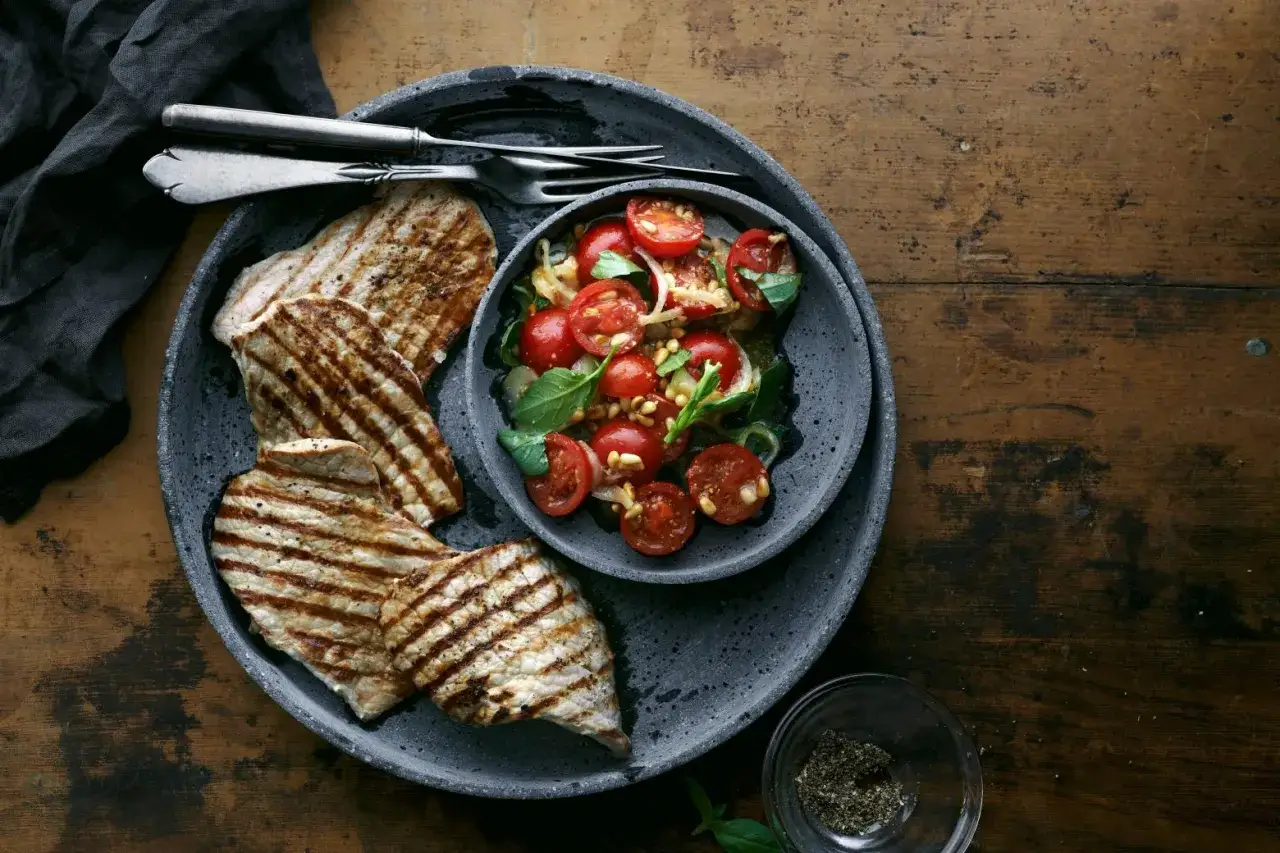 Delizioso paillard di vitello grigliato servito con un'insalata fresca di pomodorini, basilico e pinoli su un piatto scuro.