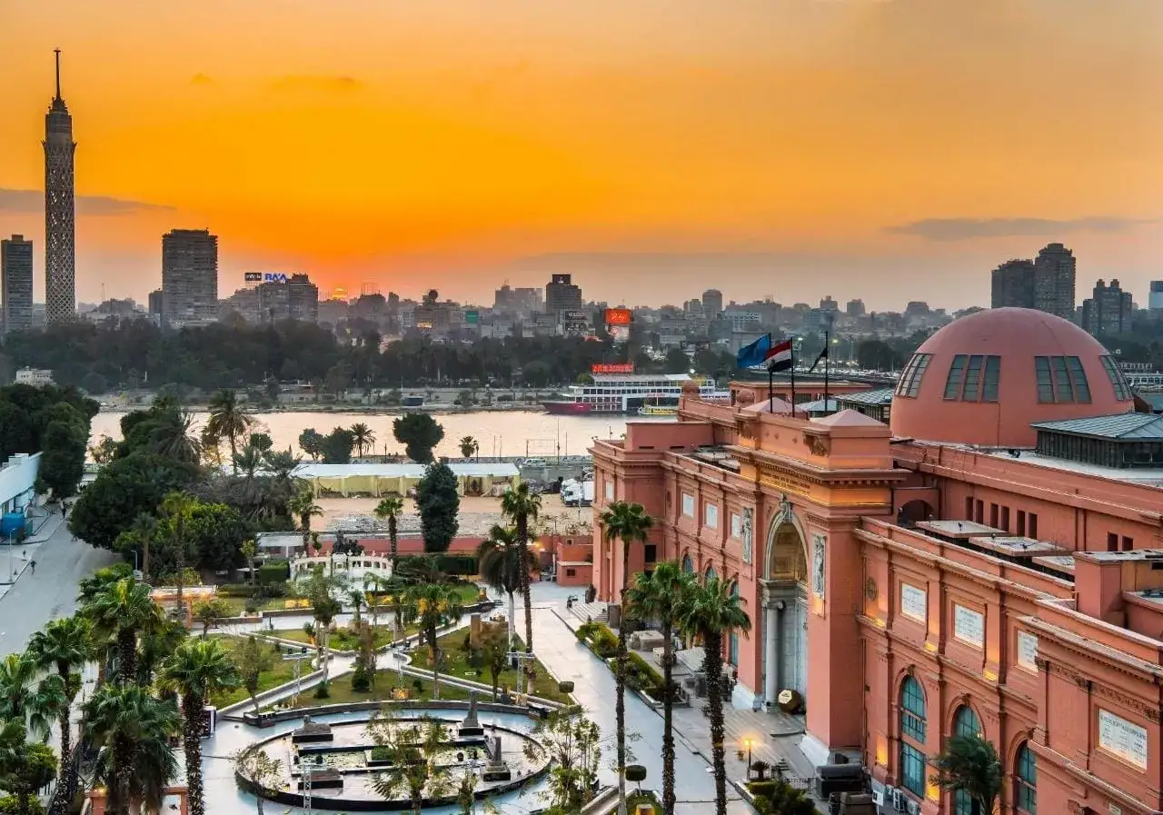Sonnenuntergang &uuml;ber Kairo. Palmen s&auml;umen den Weg zum Museum, w&auml;hrend die Skyline der Stadt im warmen Licht erstrahlt.