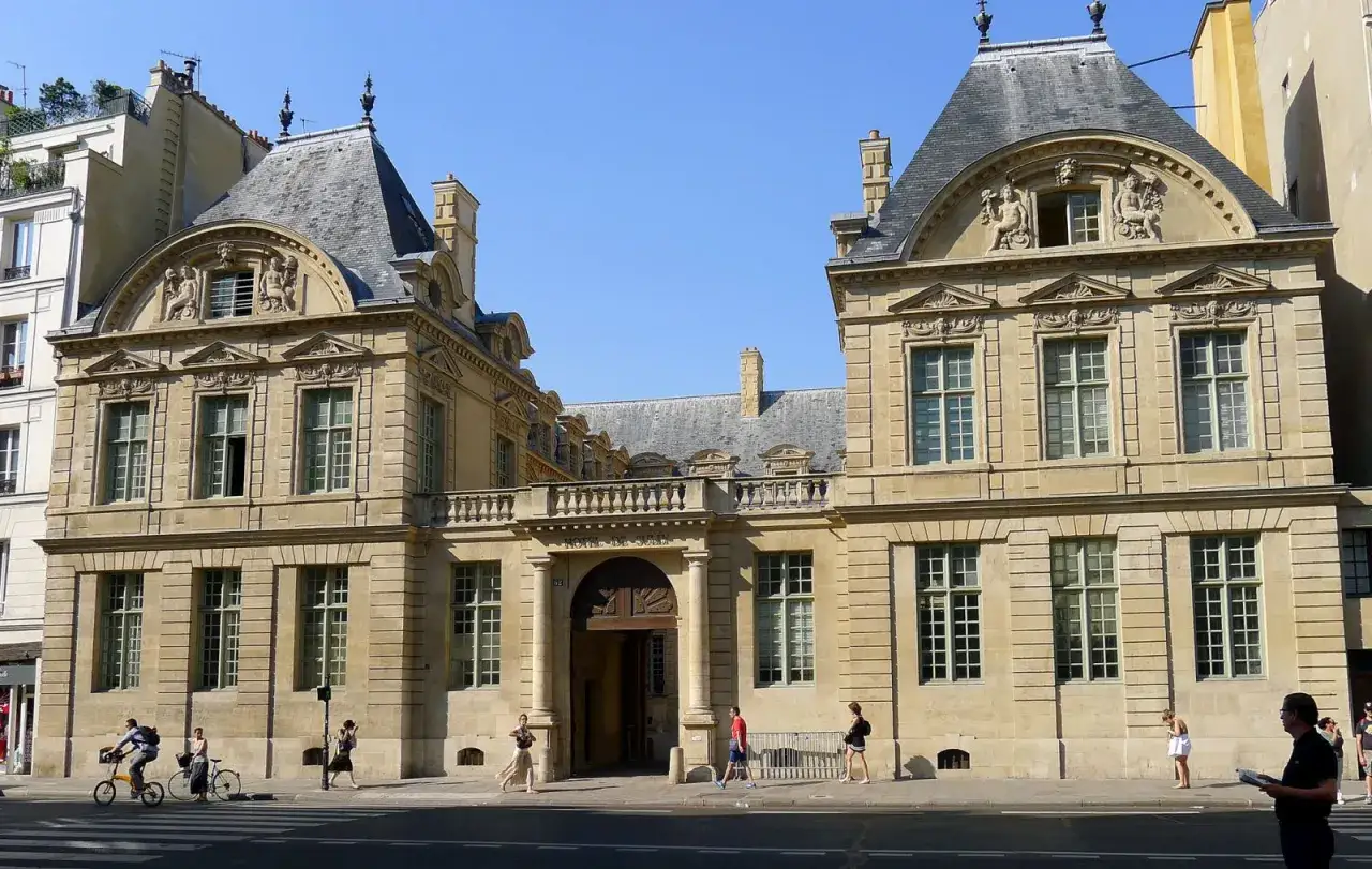 Fa&ccedil;ade d'un h&ocirc;tel particulier parisien, style baroque, avec ses sculptures et ses toits mansard&eacute;s. Des passants fl&acirc;nent sur le trottoir.
