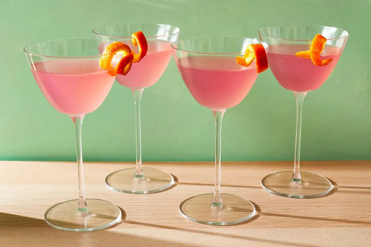 Cztery różowe drinki Cosmopolitan, każdy ozdobiony skórką pomarańczową. Idealny przepis na Cosmopolitan na letnie wieczory.