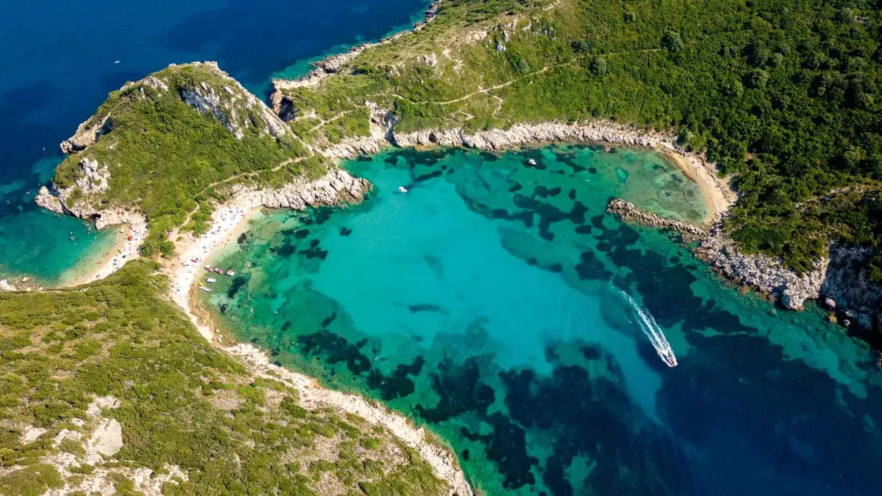 Traumhafter Korfu Strand mit türkisfarbenem Wasser, grünen Hügeln und einem Boot, das über die Wellen gleitet.