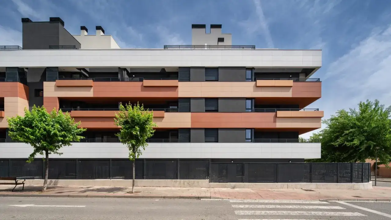 Edificio moderno con revestimiento exterior térmico en tonos tierra y blanco. Balcones volados y árboles verdes en primer plano.