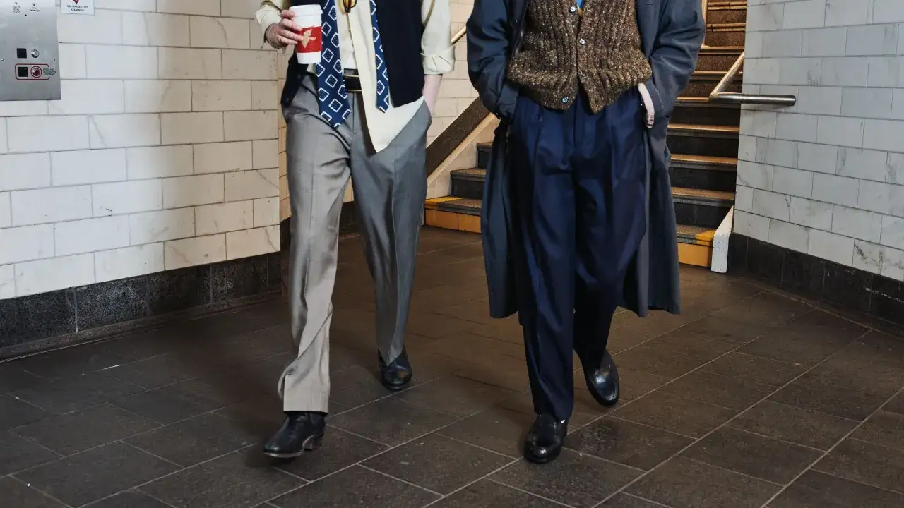 Two men walk, showcasing different trouser styles: one in grey, the other in navy.