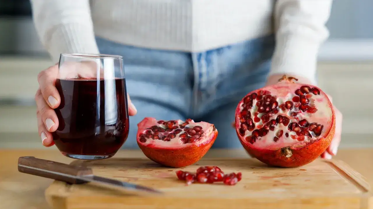 Eine Hand hält ein Glas Granatapfelsaft. Daneben liegen auf einem Holzbrett ein aufgeschnittener Granatapfel und Kerne. Granatapfel gesund!