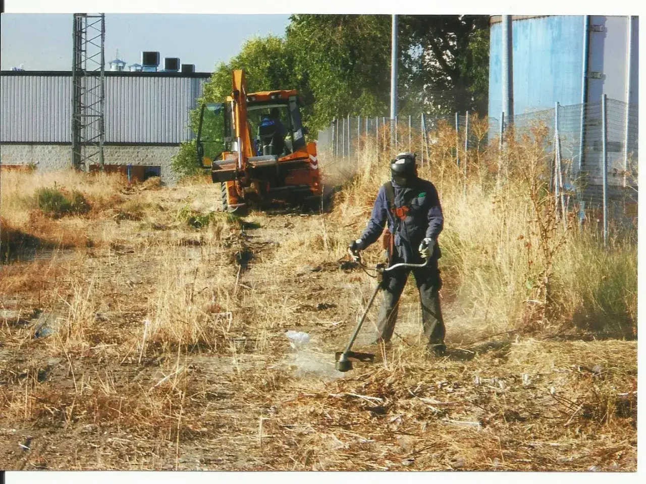Hombre con desbrozadora realiza desbroce de terreno. Una retroexcavadora naranja est&aacute; estacionada cerca.