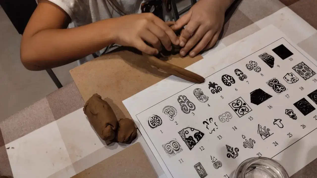 Bambino crea lavoretti sugli etruschi con argilla, ispirato da disegni su un foglio.