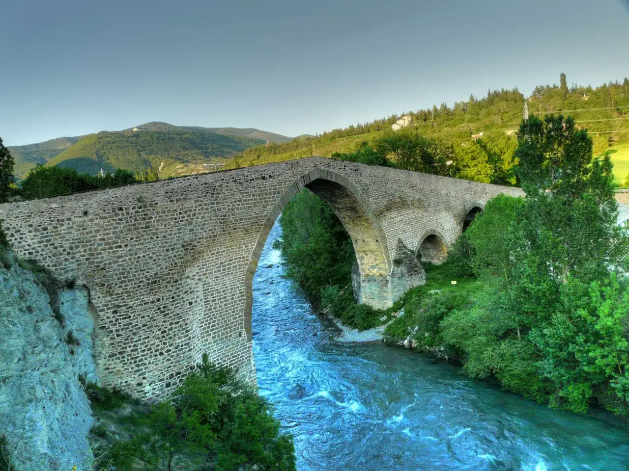 El histórico puente de San Miguel en Jaca cruza un río caudaloso rodeado de vegetación y montañas.