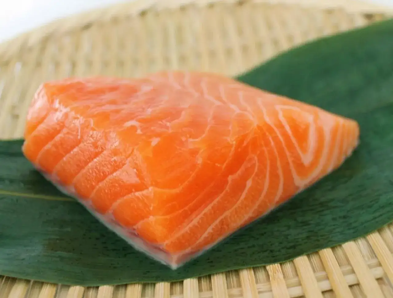 Idealny łosoś do sushi: soczysty, pomarańczowy kawałek ryby z widocznymi białymi żyłkami, ułożony na zielonym liściu na bambusowej macie.
