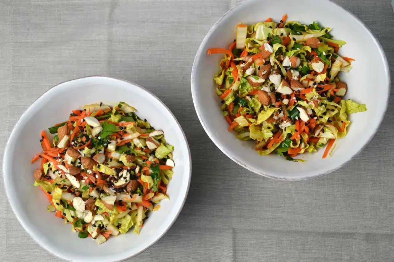 Zwei Schüsseln mit knackigem Chinakohl Salat, garniert mit geriebenen Karotten, Mandeln und schwarzen Sesam.